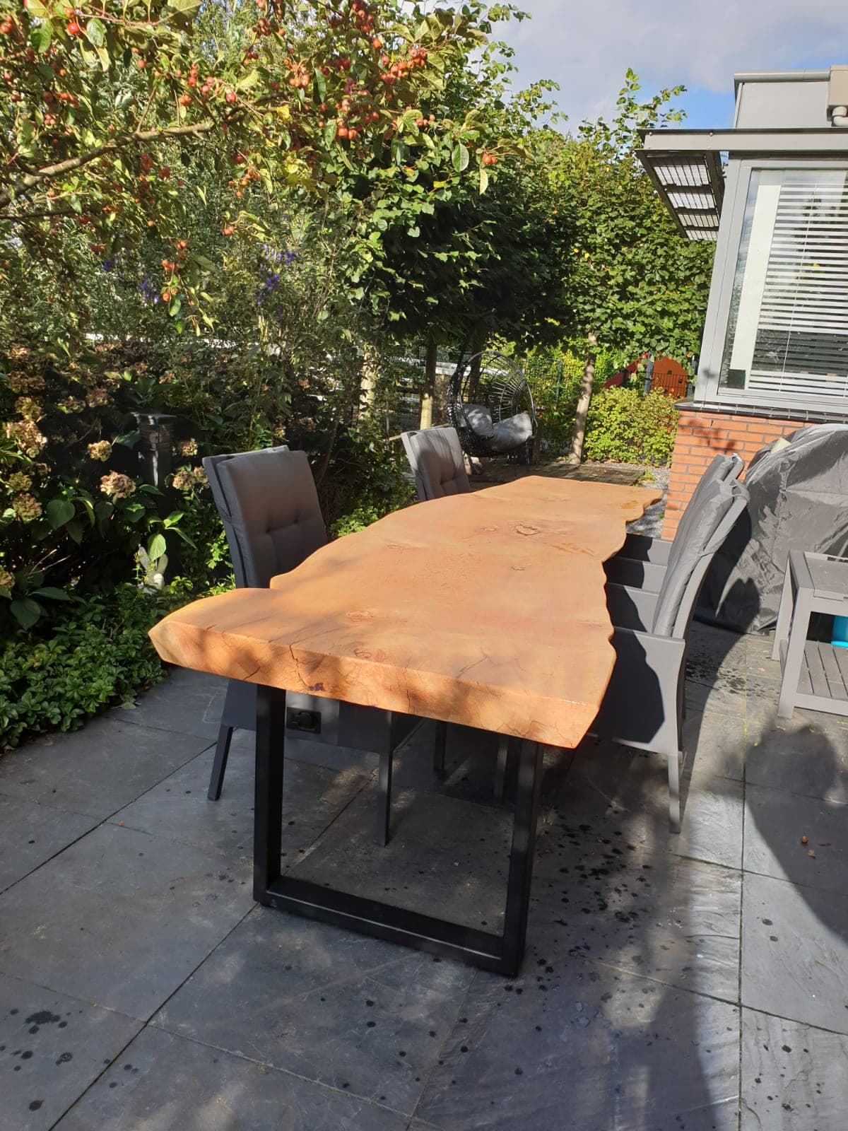 Cedar garden table with natural live edges and black U-shaped legs, surrounded by grey chairs on a terrace.