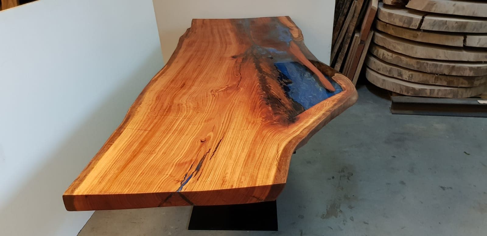Eucalyptushouten eettafel met lange roodgouden houtlijnen, blauwe epoxy langs één zijde en een natuurlijke rand in een werkplaats.