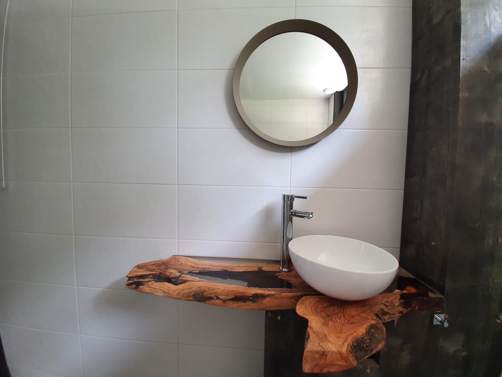 Olijfhouten hoekwastafelblad met transparante epoxy, een witte waskom en chromen kraan onder een ronde spiegel.