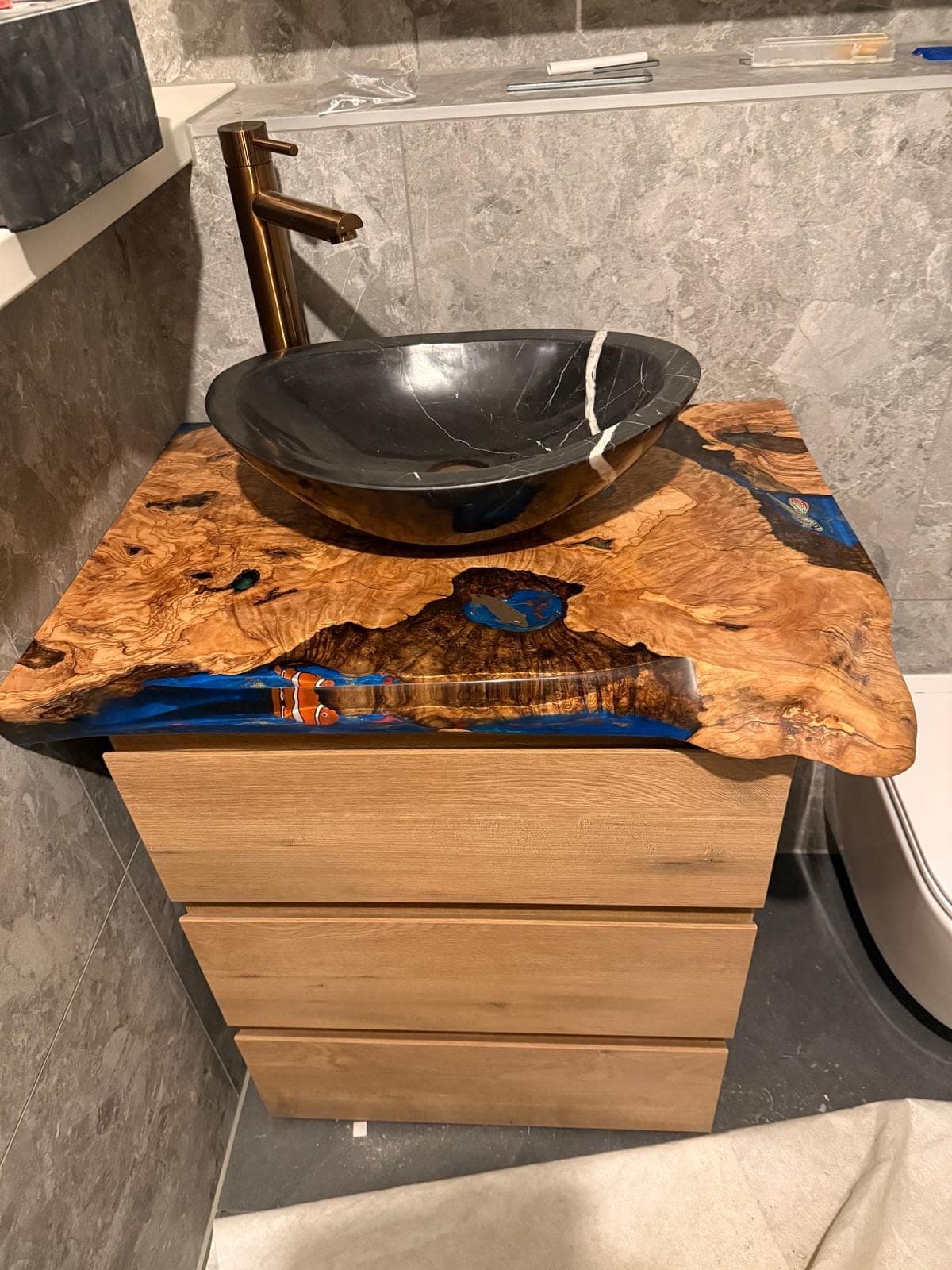 Olive wood vanity top with blue epoxy and an orange fish at the front edge, beneath a black vessel basin.