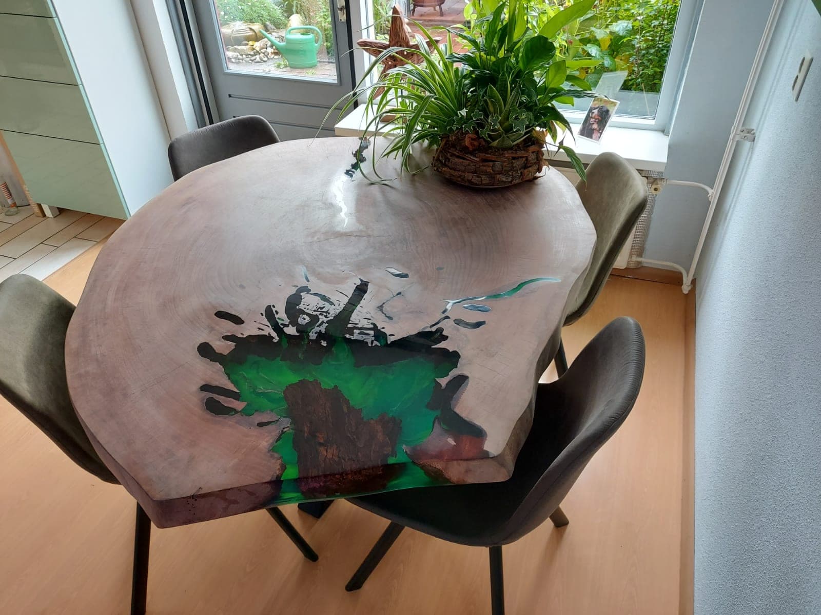 Table ronde en cèdre aux cernes visibles, avec une cavité remplie de résine verte au bord, entourée de chaises près d’une fenêtre.